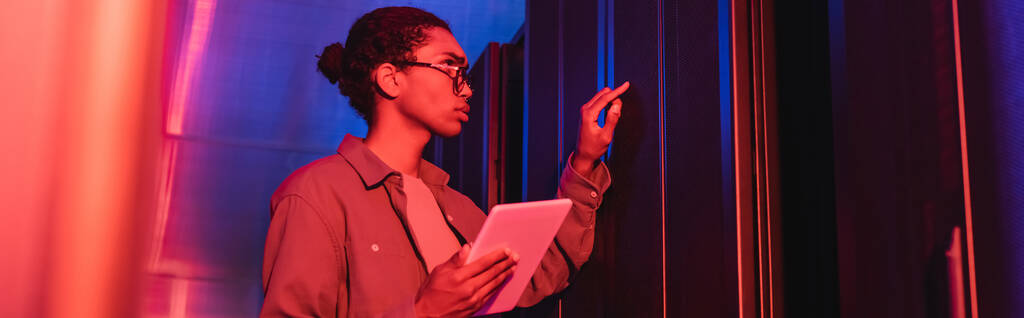 Αφρικανός αμερικανός τεχνικός με ψηφιακό tablet που ελέγχει τον server στο data center σε neon light, banner - Φωτογραφία, εικόνα