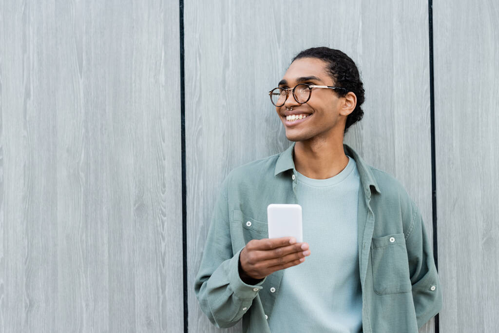 uomo afroamericano alla moda con cellulare sorridente mentre guarda lontano vicino al muro grigio - Foto, immagini