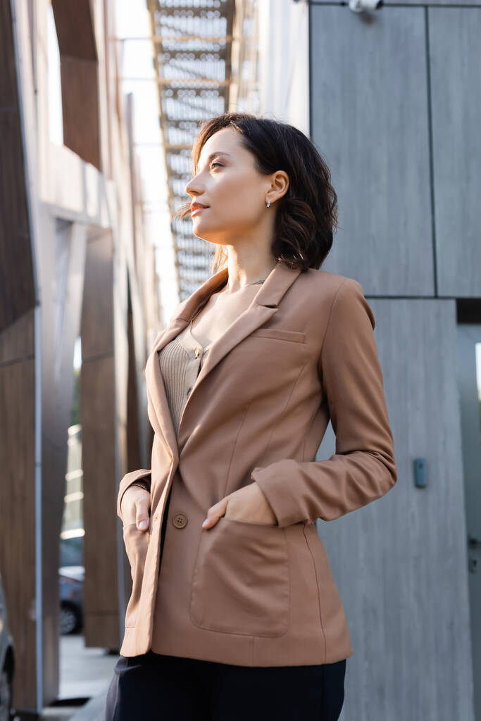 νεαρή γυναίκα κοιτάζει μακριά, ενώ στέκεται με τα χέρια στις τσέπες του μπεζ σακάκι σε εξωτερικούς χώρους - Φωτογραφία, εικόνα