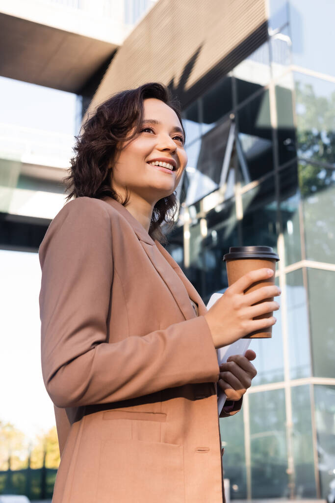 χαμηλή γωνία άποψη της κομψής γυναίκας με καφέ για να πάει χαμογελώντας, ενώ κοιτάζοντας μακριά σε εξωτερικούς χώρους - Φωτογραφία, εικόνα