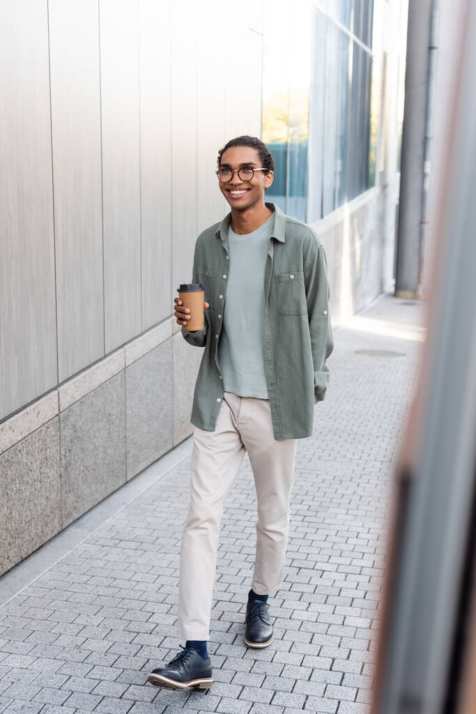 若いスタイリッシュなアフリカ系アメリカ人男性がコーヒーを飲みながら通りを歩く様子を長時間見ながら - 写真・画像
