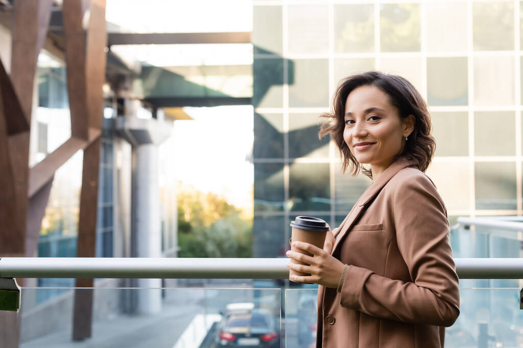 young woman with coffee to go looking at camera on balcony outdoors - Photo, Image