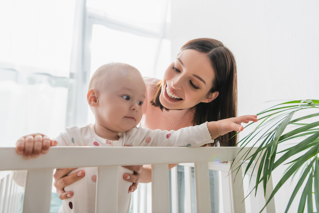 happy mother holding baby boy touching plant while standing in crib - Photo, Image