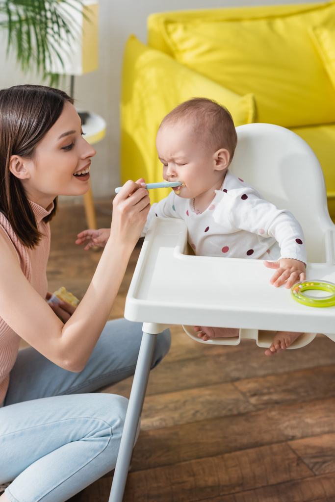 happy woman feeding little son sitting in baby chair with closed eyes - Photo, Image