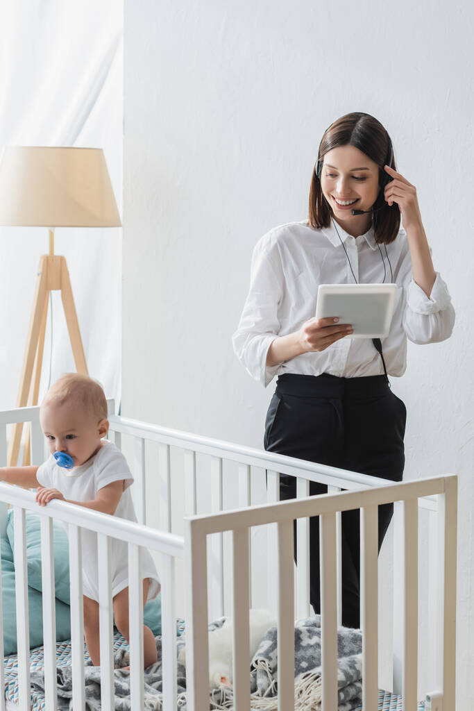 Lächelnde Frau mit digitalem Tablet arbeitet im Headset neben Kleinkind im Kinderbett - Foto, Bild