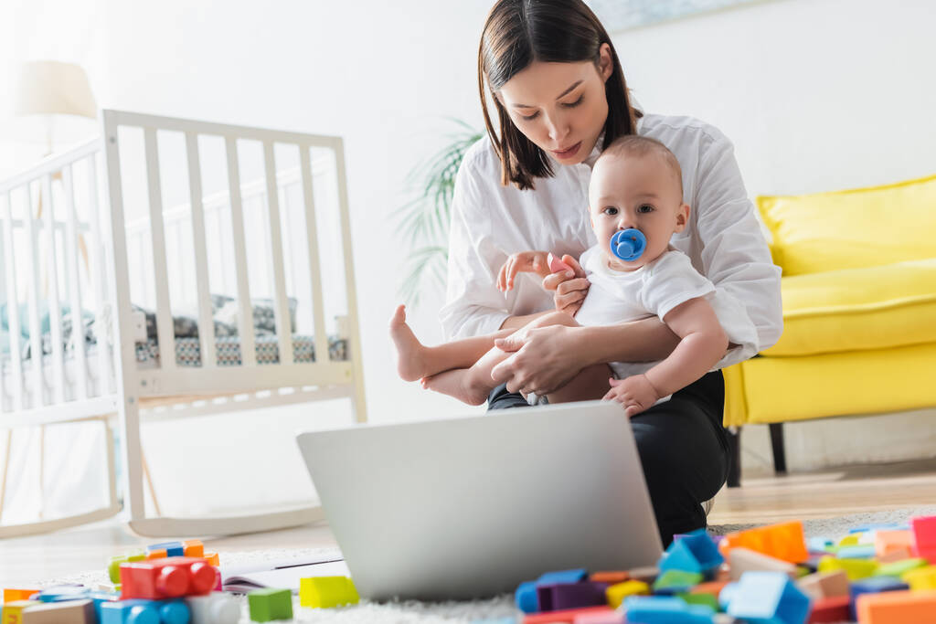 mother holding little kid while sitting on floor near laptop and blurred building blocks - Photo, Image
