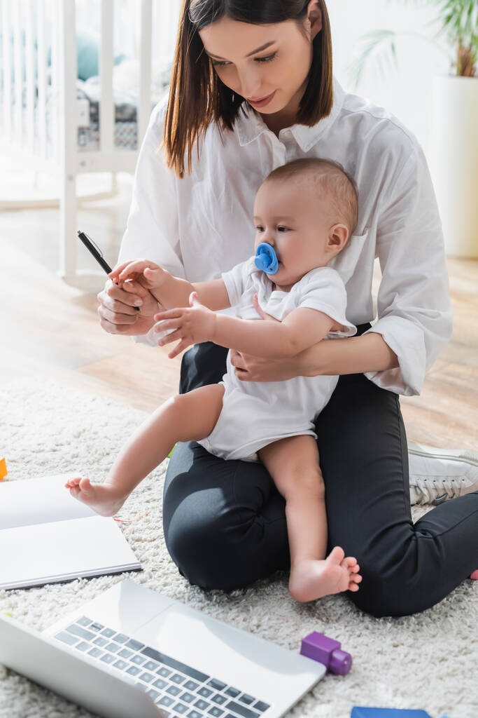 mulher mostrando caneta para pequeno filho enquanto sentado no chão perto notebook - Foto, Imagem