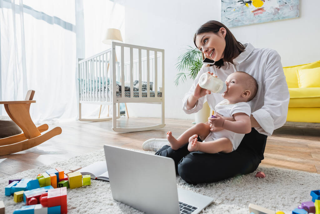 smiling woman talking on smartphone while feeding son on floor near laptop and building blocks - Photo, Image