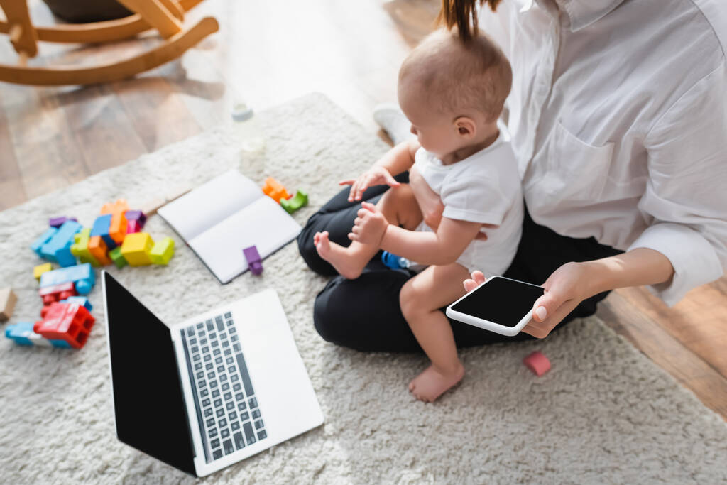 wysoki kąt widzenia kobiety z małym synem i smartfonem siedzi na podłodze w pobliżu laptopa i bloków - Zdjęcie, obraz