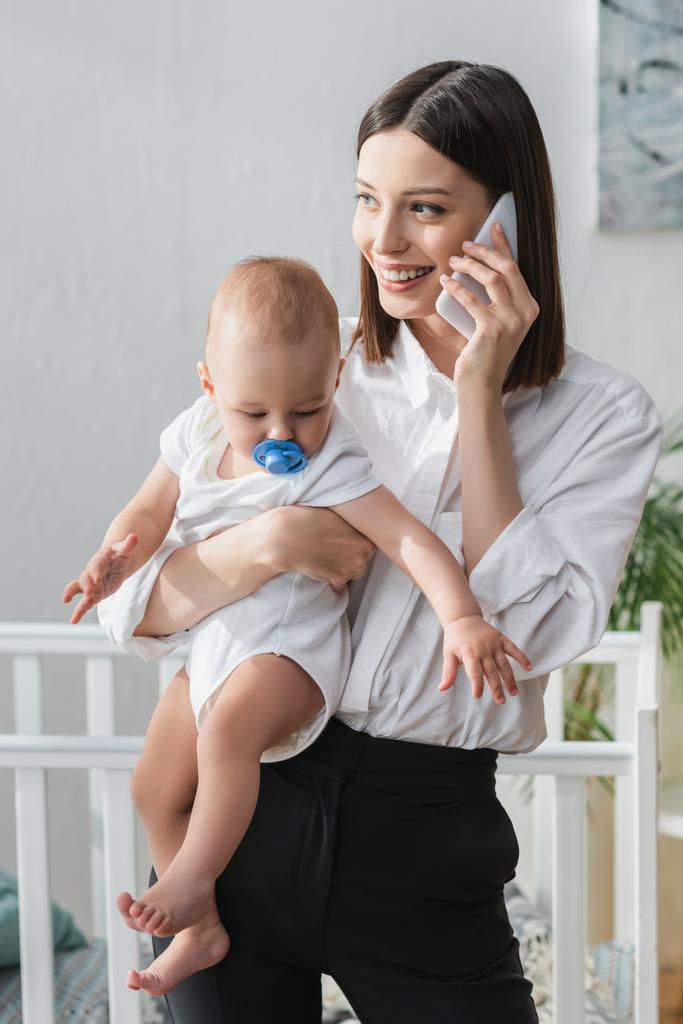 mujer sonriente hablando en el teléfono inteligente mientras sostiene al bebé en las manos - Foto, Imagen