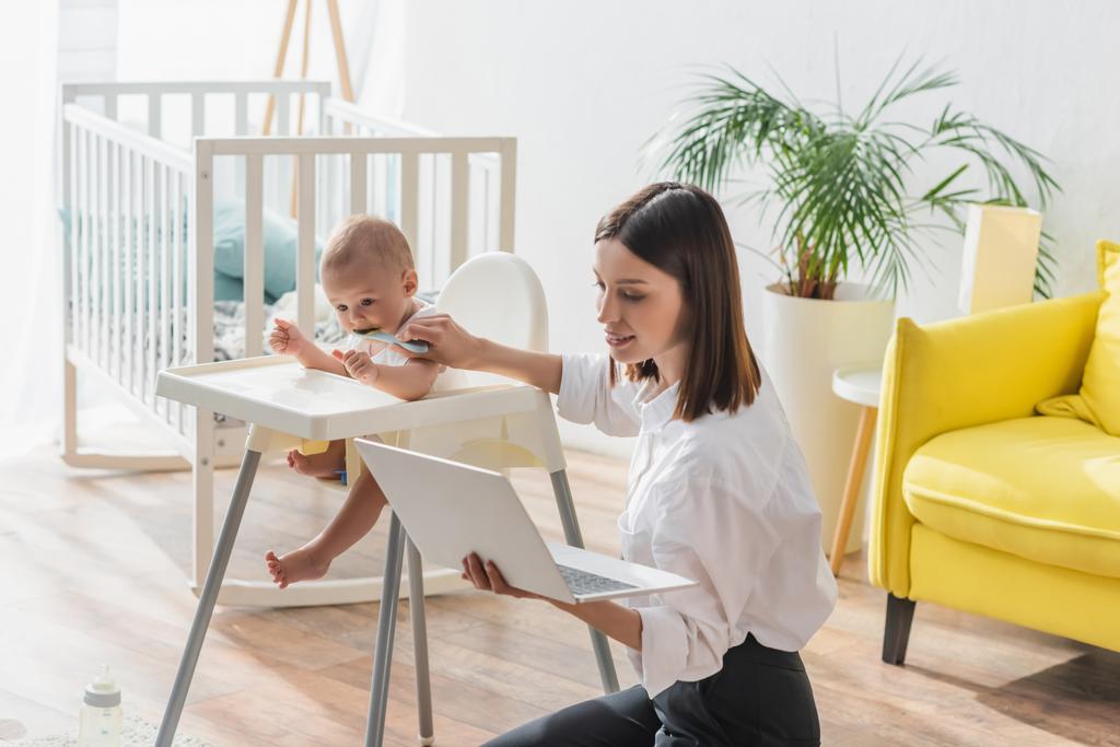 brunetka kobieta siedzi na podłodze z laptopem podczas karmienia syn w dziecko krzesło - Zdjęcie, obraz