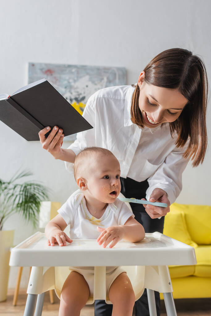 mujer alegre con cuaderno de alimentación pequeño hijo con puré en casa - Foto, Imagen