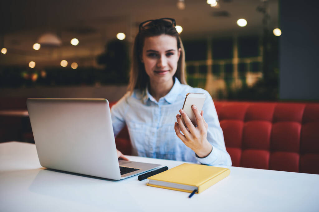 Boldog női távmunkás jegyzettömbbel és modern laptoppal az asztalnál ülve a könnyű büfében és okostelefonnal a kamerába nézve - Fotó, kép