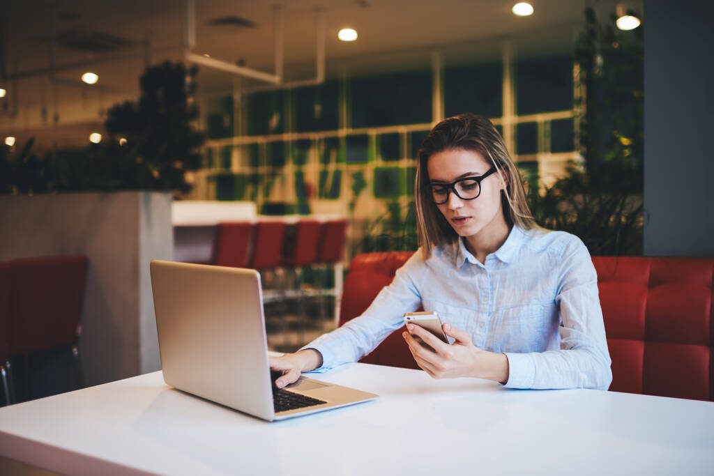 Huomaavainen nainen freelancer silmälaseissa ja kevyessä paidassa selaamassa kannettavaa tietokonetta ja älypuhelimella työskennellessään nykyaikaisessa kahvilassa päivällä - Valokuva, kuva