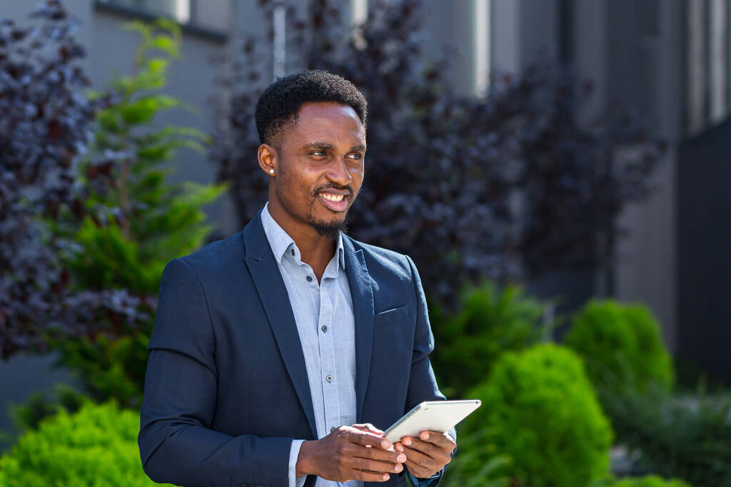 Fiatal afro-amerikai üzletember, hivatalos öltönyben, táblagéppel a kezében a háttérben, modern irodaházon kívül. A férfi okostelefont vagy mobiltelefont használ a város utcájában - Fotó, kép