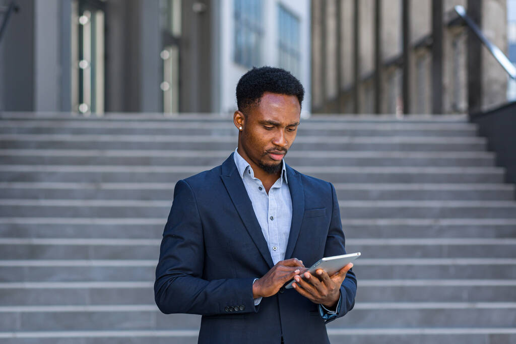 Ein junger afrikanisch-amerikanischer Geschäftsmann im formellen Businessanzug steht mit Tablet in der Hand vor einem modernen Bürogebäude im Hintergrund. Mann benutzt Smartphone oder Handy im Freien - Foto, Bild