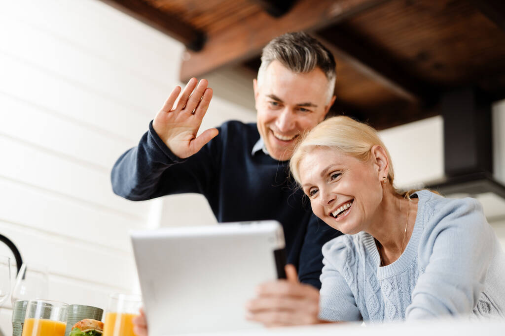 Χαμογελώντας μεσήλικες ζευγάρι σε μια βιντεοκλήση μέσω ψηφιακής tablet από το σπίτι, wawing hand - Φωτογραφία, εικόνα