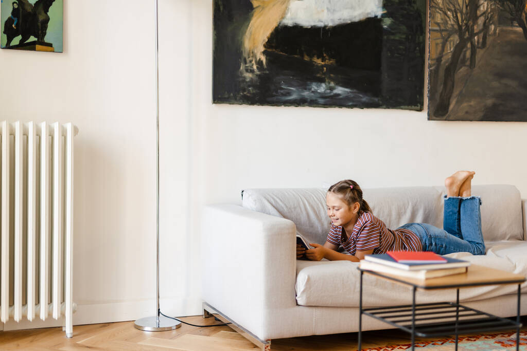 Chica preadolescente blanca sonriendo y usando el teléfono móvil mientras está acostada en el sofá en casa - Foto, Imagen