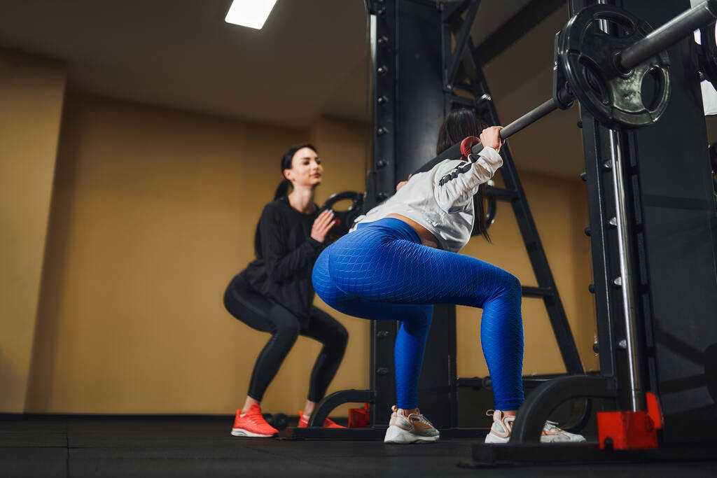 Junge kaukasische Frau trainiert Beine mit Langhantel Im Fitnessstudio mit Trainer. Persönliche Trainerin - Foto, Bild
