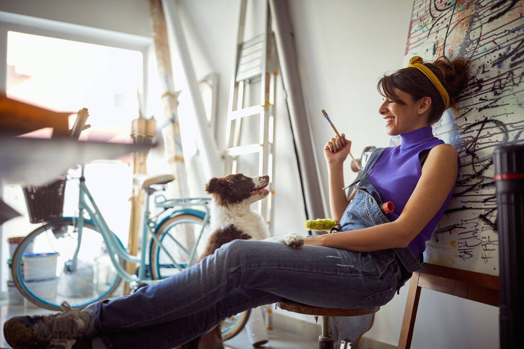 Una joven artista que se divierte con su perro en un ambiente agradable en el estudio. Arte, pintura, amistad, estudio - Foto, imagen