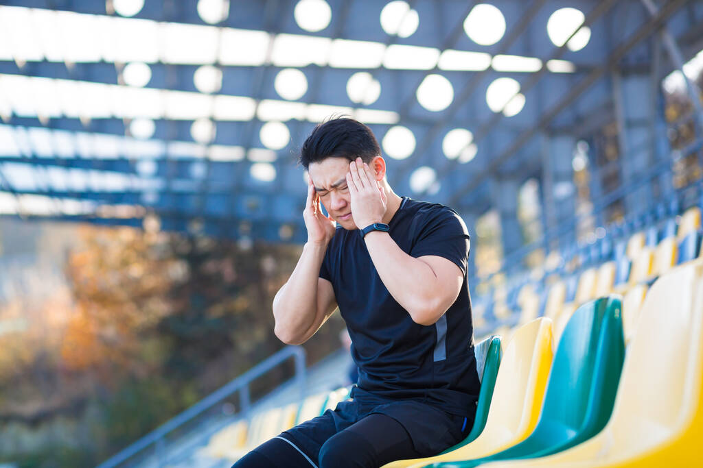 Fáradt férfi sportoló pihen, ázsiai tartja a fejét, van egy súlyos fejfájás futás után - Fotó, kép