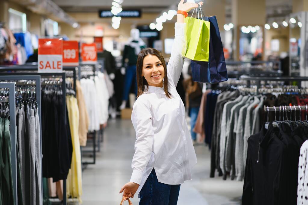 venta, ropa, compras, moda y concepto de personas - mujer joven feliz elegir entre dos camisas en el centro comercial o tienda de ropa
 - Foto, imagen