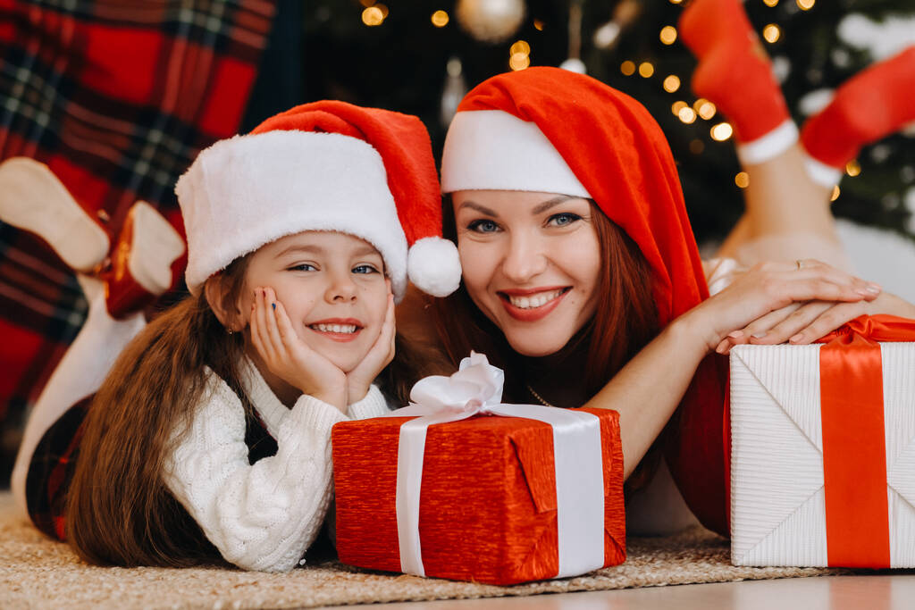 サンタクロースの帽子の幸せな女の子と彼女のお母さんは彼らの手に贈り物で笑っています. - 写真・画像