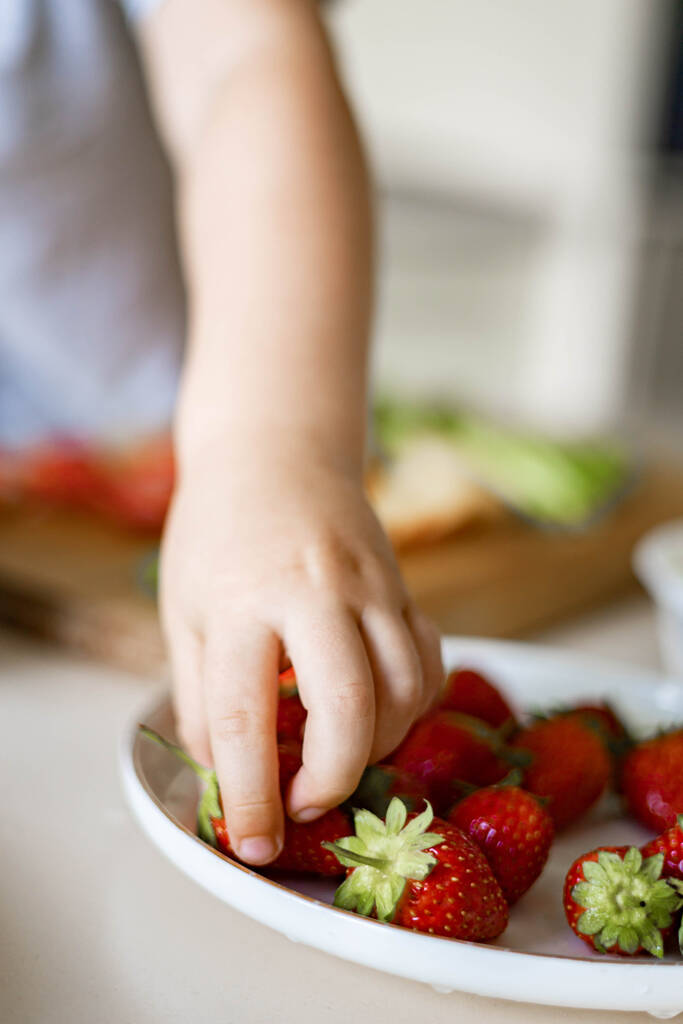 Рука крупным планом милый малыш берет свежую клубнику красной ягоды с тарелки сезонный урожай - Фото, изображение