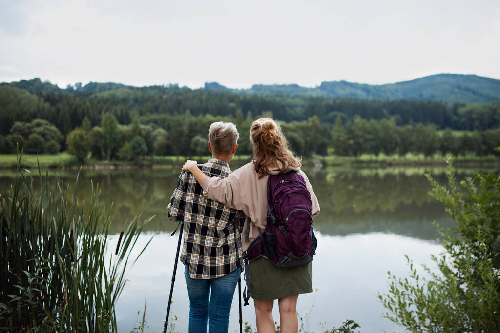 自然の中で湖の外に立っているときに大人の娘と一緒に受け入れるシニア母親の背面ビュー - 写真・画像