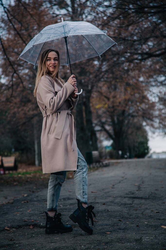 Paltolu genç, narin, güzel bir kız parkta şeffaf bir şemsiyeyle yağmurda yürüyor. - Fotoğraf, Görsel