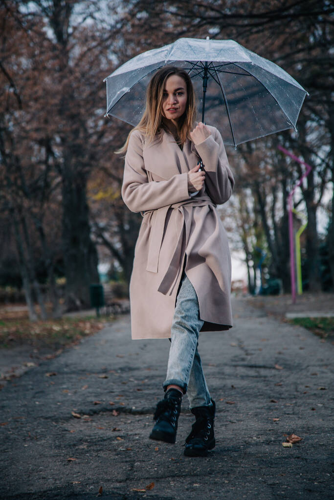 Egy fiatal, karcsú, gyönyörű lány kabátban sétál az esőben egy átlátszó esernyővel a parkban. - Fotó, kép