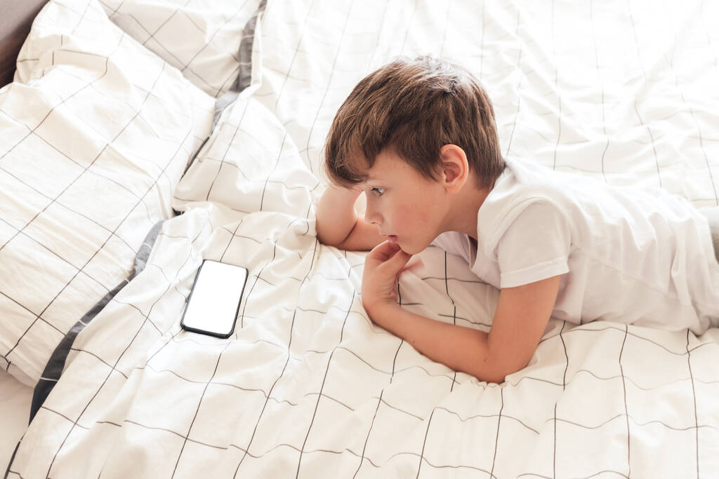Dítě sedí uvolněně v posteli a hraje na smartphonu - Fotografie, Obrázek