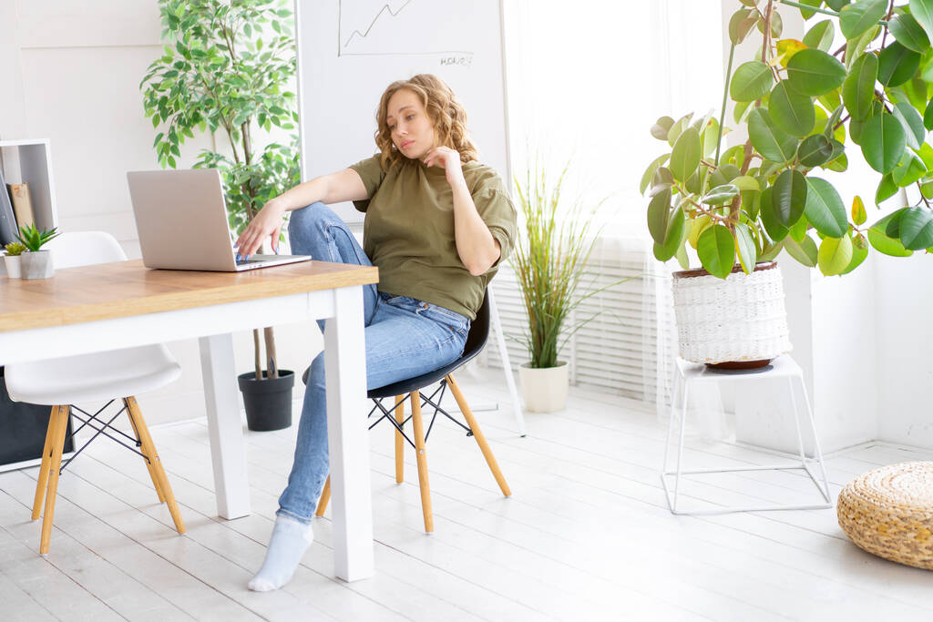 Geschäftsfrau mit Laptop sitzend Schreibtisch weiß Büroeinrichtung mit Zimmerpflanze aussehende Geschäftsleute Geschäftsmann Online, Jung und erfolgreich Gekleidet grünes Hemd blaue Jeans barfuß entspannend  - Foto, Bild
