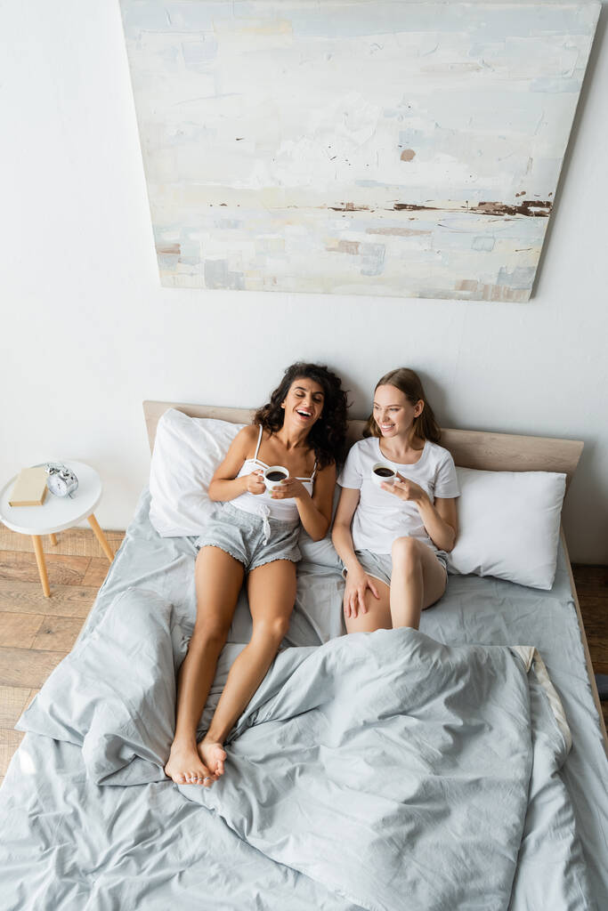 вид под высоким углом на счастливую лесбийскую пару, держащую чашки кофе в постели - Фото, изображение