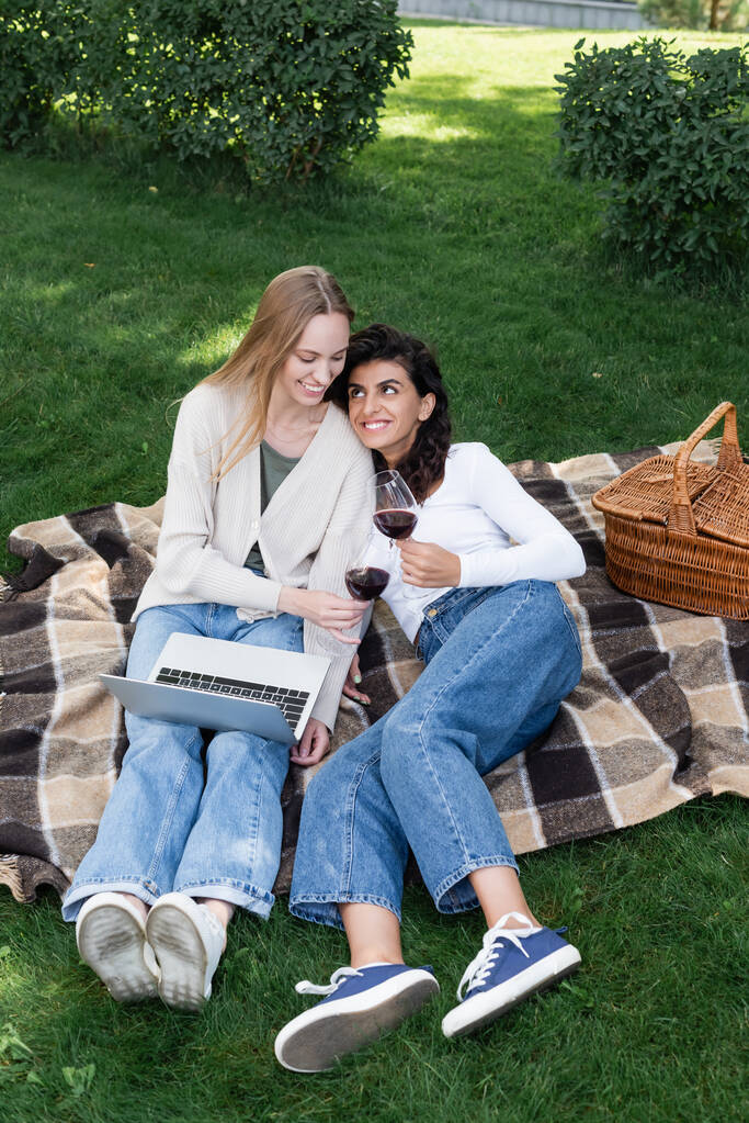 ピクニック中にノートパソコンの近くで赤ワインのグラスを持っている幸せな女性  - 写真・画像