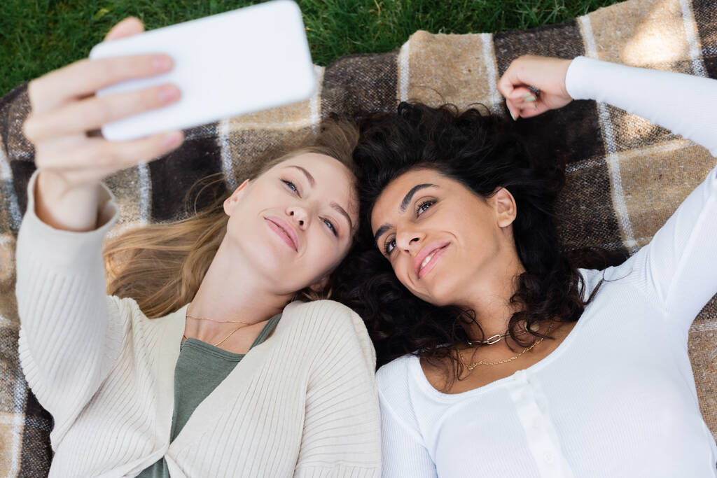 vysoký úhel pohledu na šťastné ženy brát selfie, zatímco leží na dece v parku  - Fotografie, Obrázek