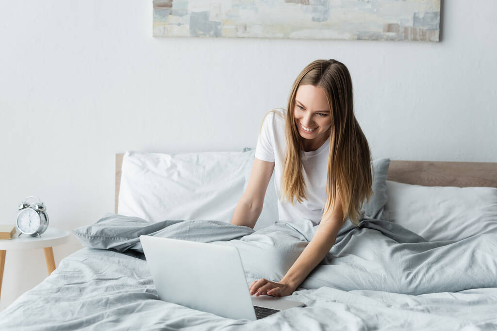 gelukkig jong freelancer met behulp van laptop terwijl zitten in bed - Foto, afbeelding