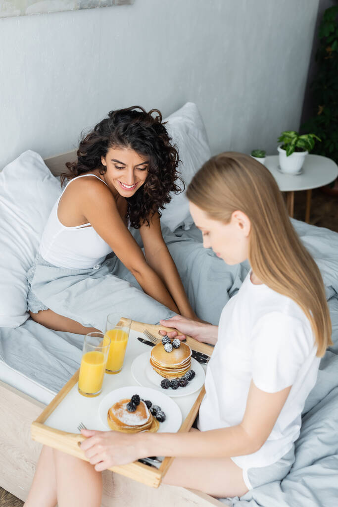 tevreden jonge vrouw houden lade met smakelijk ontbijt in de buurt krullend vriendin in bed - Foto, afbeelding