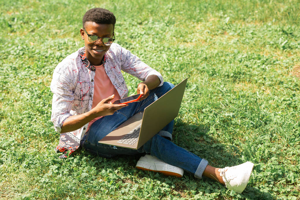 Αφρικανός φοιτητής κάθεται στο γρασίδι με φορητό υπολογιστή και τηλέφωνο - Φωτογραφία, εικόνα