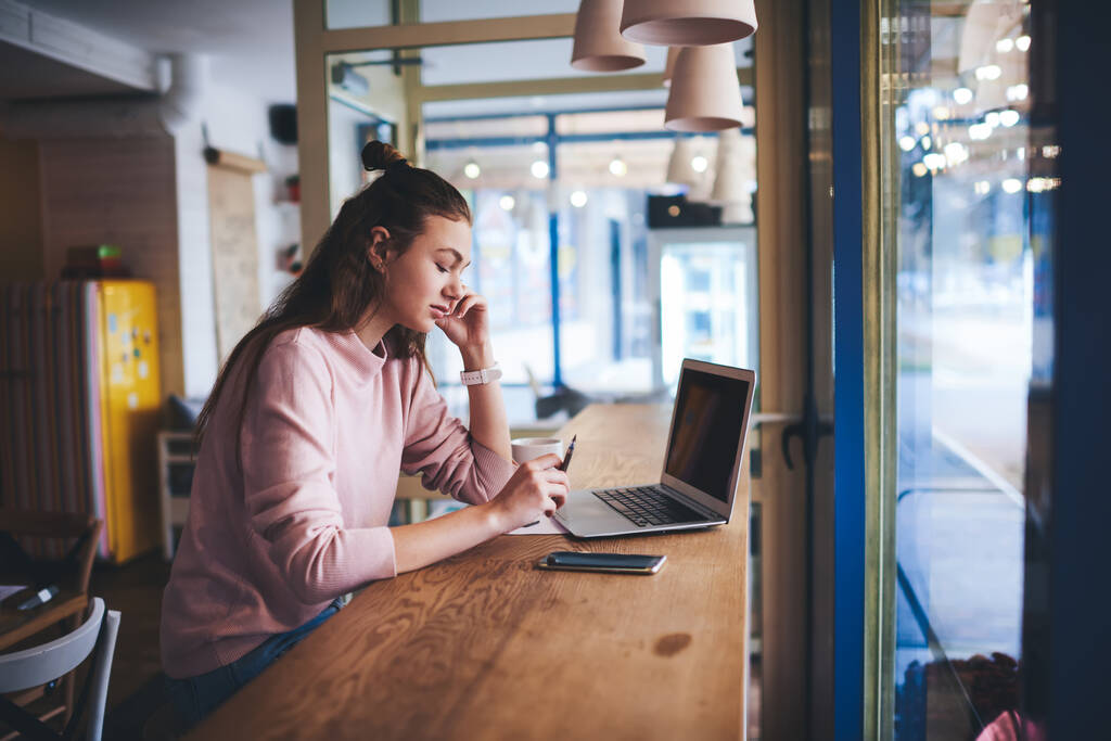 Seitenansicht einer nachdenklichen Dame in lässiger Kleidung, die sich Notizen ansieht, während sie tagsüber im gemütlichen Café ferngesteuert an Gadgets arbeitet - Foto, Bild