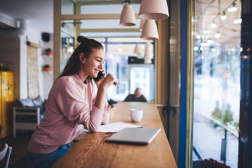 Seitenansicht einer jungen lächelnden Freiberuflerin, die am Tisch mit Laptop und Tasse Getränk auf dem Handy spricht, während sie von einem Café aus ferngesteuert arbeitet - Foto, Bild