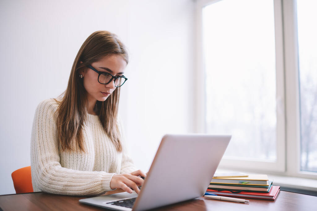 Gözlüklü ve beyaz kazaklı meşgul genç bir kadın, hafif çalışma alanındaki ahşap masada çalışırken taşınabilir laptopta bilgileri kontrol ediyor. - Fotoğraf, Görsel