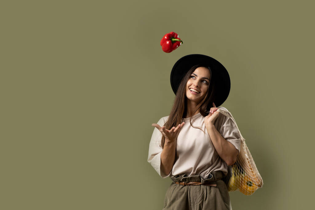 Smiling brunette young woman in a beige t-shirt and a hat holding a reusable mesh bag with fresh vegetables and fruits on a shoulder and throwing a red pepper in a air. Zero waste concept. - Photo, Image