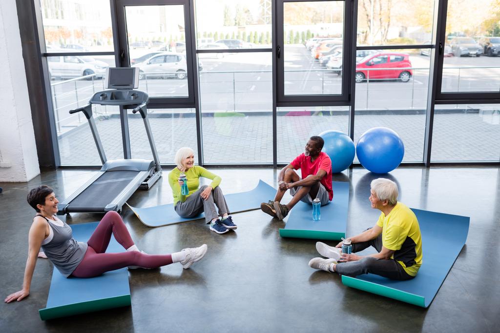 Happy interracial senior people holding sports bottles while sitting on fitness mats in gym  - Photo, Image