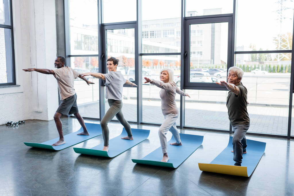 Multicultural elderly people standing in warrior pose on mats in sports center  - Photo, Image