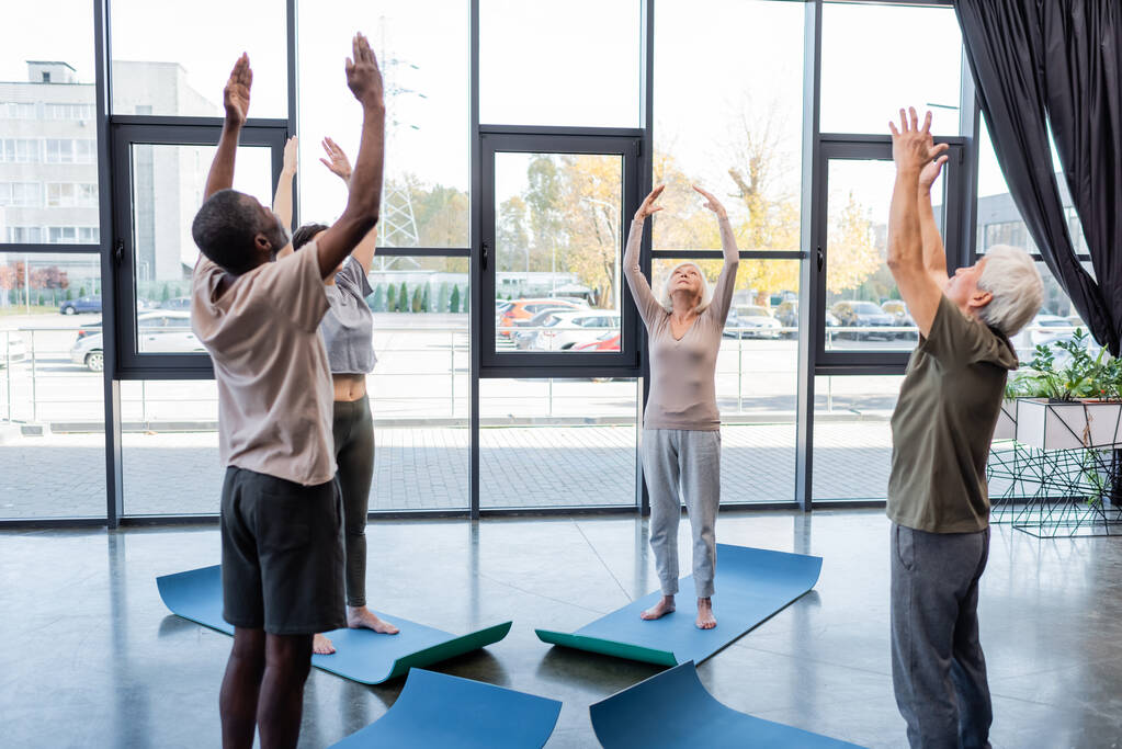 Senior woman raising hands while practicing yoga near multiethnic friends in sports center - Photo, Image