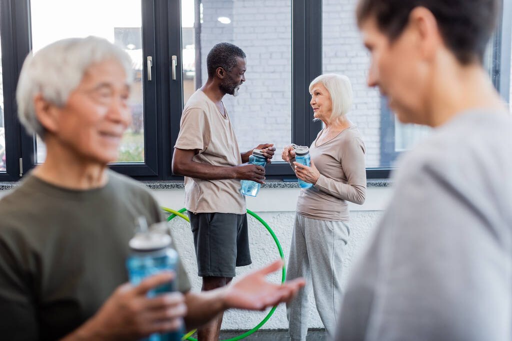 Multiethnische Senioren halten Sportflaschen in der Hand, während sie im Fitnessstudio reden  - Foto, Bild