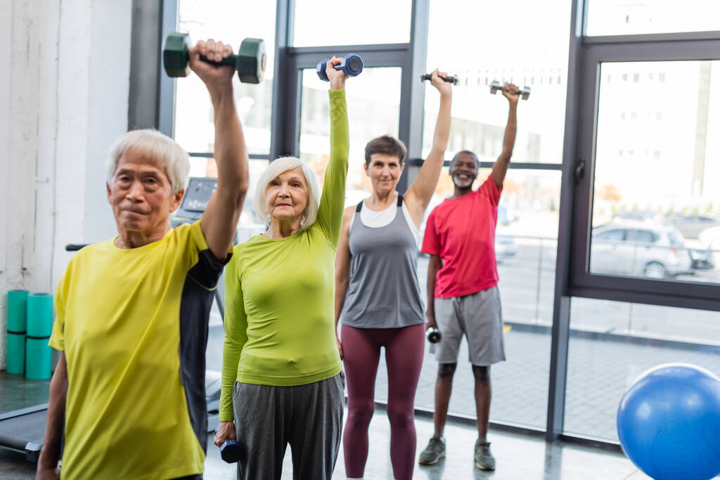 Grupo de personas mayores interraciales haciendo ejercicio con mancuernas en un centro deportivo  - Foto, Imagen