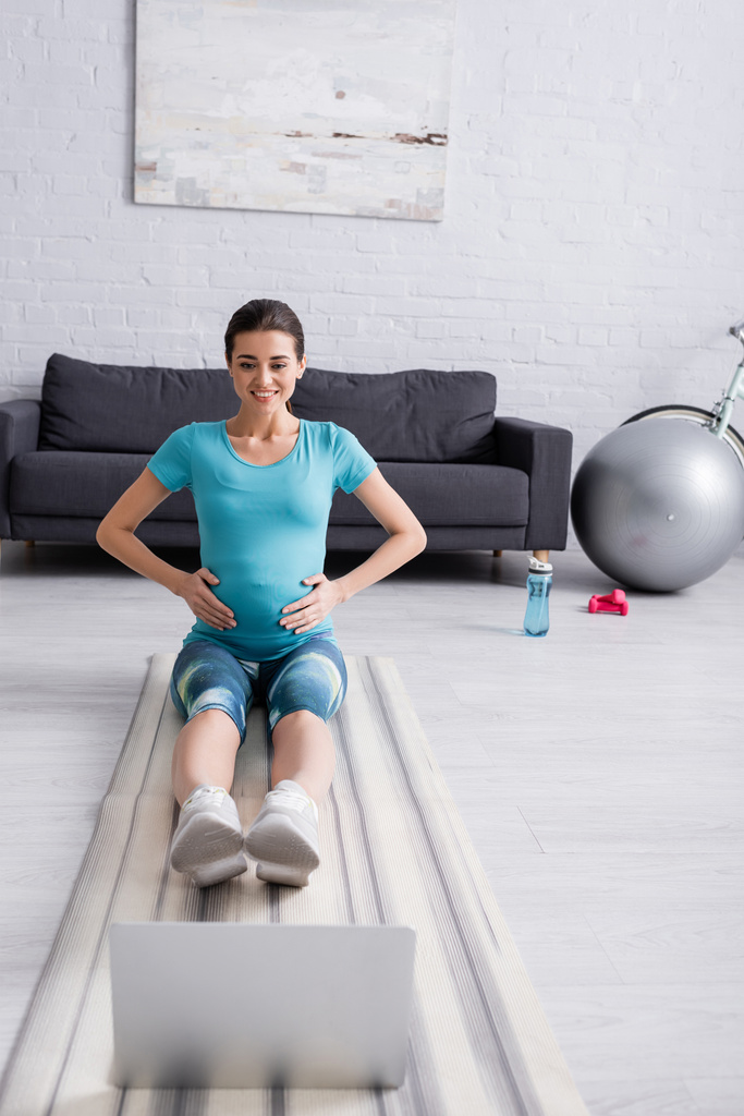 mujer embarazada sonriente en ropa deportiva ejercicio cerca de la computadora portátil en la sala de estar  - Foto, imagen