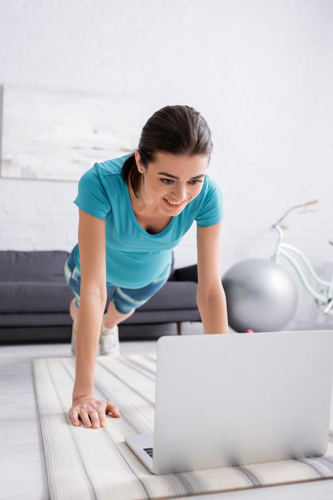 radosna ciężarna kobieta w odzieży sportowej ćwicząca w pobliżu laptopa w salonie  - Zdjęcie, obraz